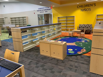 Bright yellow walls, a gray rug, a small area with an orange rug, an area with a blue rug, and book shelving around a rug with a design that looks like a river with a rope bridge across the river, surrounded by grass, rocks, and cattails. By one end of the rug is a play kitchen area. The other end has a wooden table with two wooden chairs. The teen area with shelving, a green rug, and a table with chairs is in the background.
