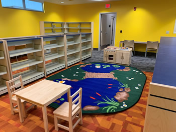 Bright yellow walls, orange rug, and book shelving around a rug with a design that looks like a river with a rope bridge across the river, surrounded by grass, rocks, and cattails. By one end of the rug is a play kitchen area. The other end has a wooden table with two wooden chairs.