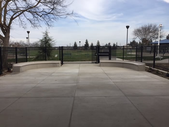 A large cement area surrounded by a black steel fence. There are two long, curved cement seating areas. One to the left and one to the right at the back of the cement area near the fence. In the background there is a large tree, a grassy area, and several smaller trees in the distance. 