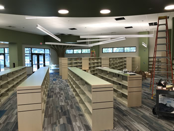 Seven sections of shelving in the Children's area of the library on gray and blue carpeting. The walls in the background are green, with a white ceiling. There are windows and a double glass door in the background.