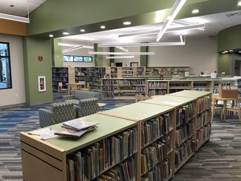 At the bottom of the picture there is a long bookshelf filled with children's books and there is a lot more shelving across the room. Next to that there are two child-sized chairs, with only the back and a small part of the side visible. The carpet is blue, gray, tan, and white in a varigated pattern. The walls are tan with green accents.