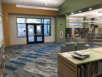 The corner of a shelving section and two child-sized chairs are in the foreground. There are several sections of shelving across the room. The carpet is blue, gray, tan, and white in a varigated pattern. The walls are tan with orange and green highlights. There is a double glass door with windows around it to the back of the area.