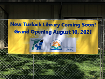 Large sign with a yellow background hanging on a chainlink fence. Words: New Turlock Library coming soon! Grand Opening August 10, 2021. 