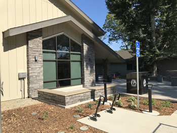 A front view of the main entrance to the Turlock Library with the black bookdrop in the center of the picture. The front sections of the library, with tan walls, some stone bricks near the entrance, and black windows.