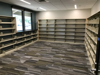 The interior area of the Friends of the Turlock Public Library bookstore. There is shelving along all the walls. The wall to the left is green and has a window in it. The other walls are tan. The carpet is gray, tan, and white.