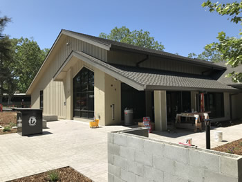 A side view of the main entrance to the Turlock Library with the black bookdrop to the left in the picture. There is a small cinderblock wall to the right in the picture. The front section of the library, with tan walls and black windows can be seen in the center of the photo with blue sky and trees above.