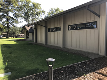 The back side of the library with windows in the tan walls. There is a small walkway in front of the walls. There is a large grass covered area with some trees in the distance.