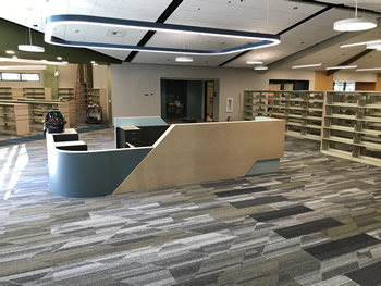 The service desk in the main area of the library, with green and tan sides for the desk. There is shelving to the right and in the background to the left. There is a rectangular light structure, with curved corners, hanging over the desk. The ceiling is white. The carpet is gray, tan, and white.